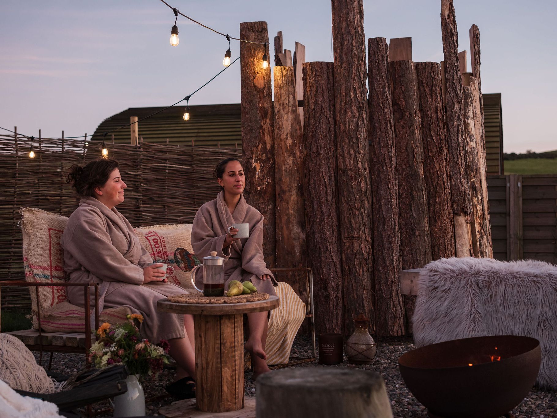 Two women in robes relaxing outdoors at a wild spa with sauna, contrast therapy, string lights, rustic wood fencing, and a fire pit.