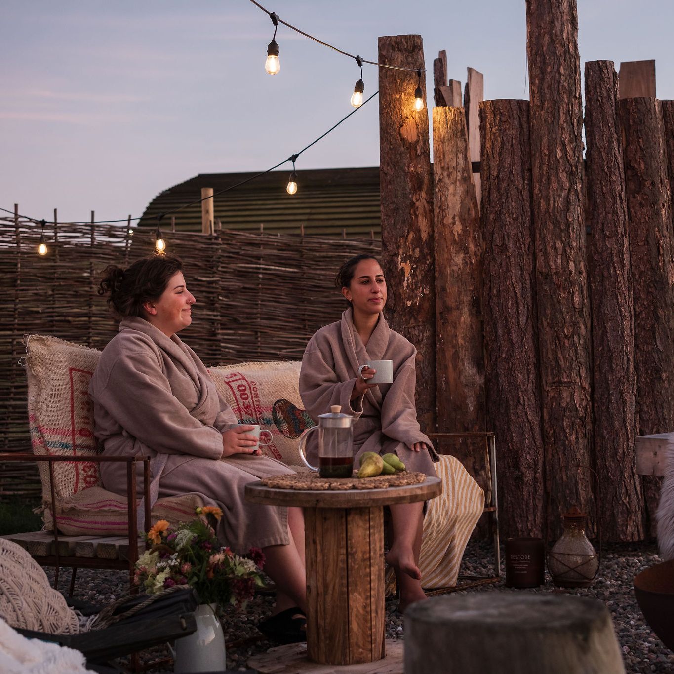 Two women in robes relaxing outdoors at a wild spa with sauna, contrast therapy, string lights, rustic wood fencing, and a fire pit.
