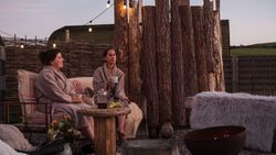 Two women in robes relaxing outdoors at a wild spa with sauna, contrast therapy, string lights, rustic wood fencing, and a fire pit.