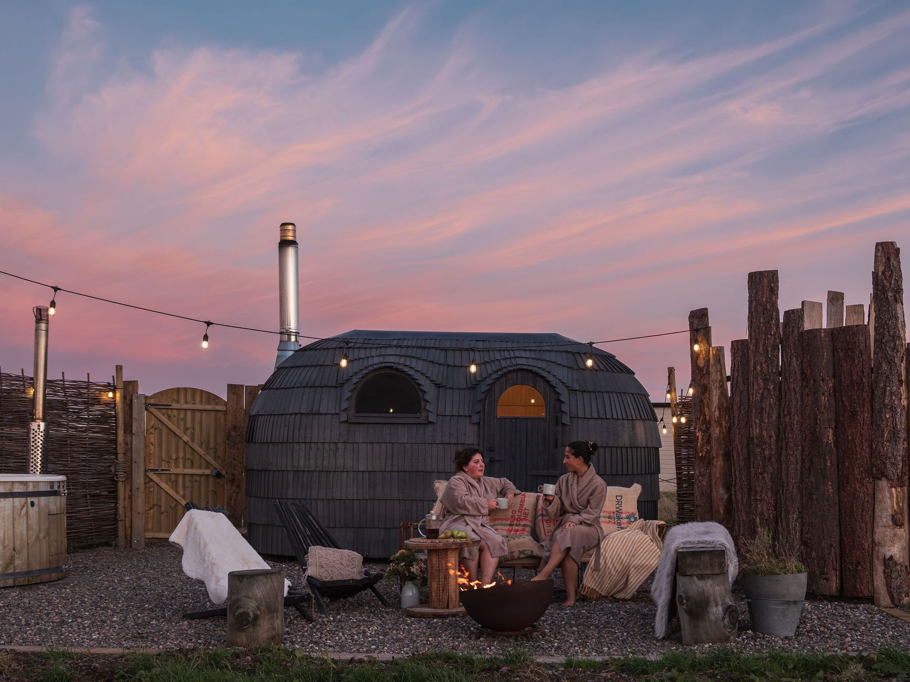 Two people in robes relax by a fire pit at an outdoor spa with a wooden hot tub and a unique Iglusauna  at sunset.