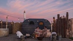 Two people in robes relax by a fire pit at an outdoor spa with a wooden hot tub and a unique Iglusauna  at sunset.