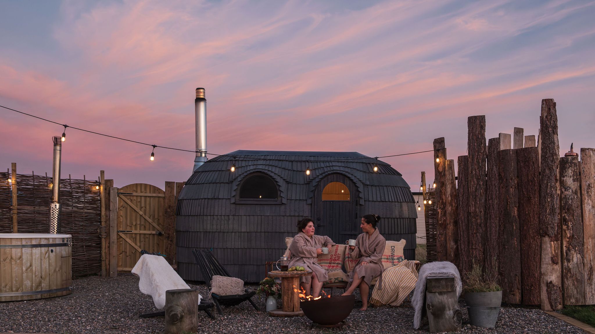 Two people in robes relax by a fire pit at an outdoor spa with a wooden hot tub and a unique Iglusauna  at sunset.