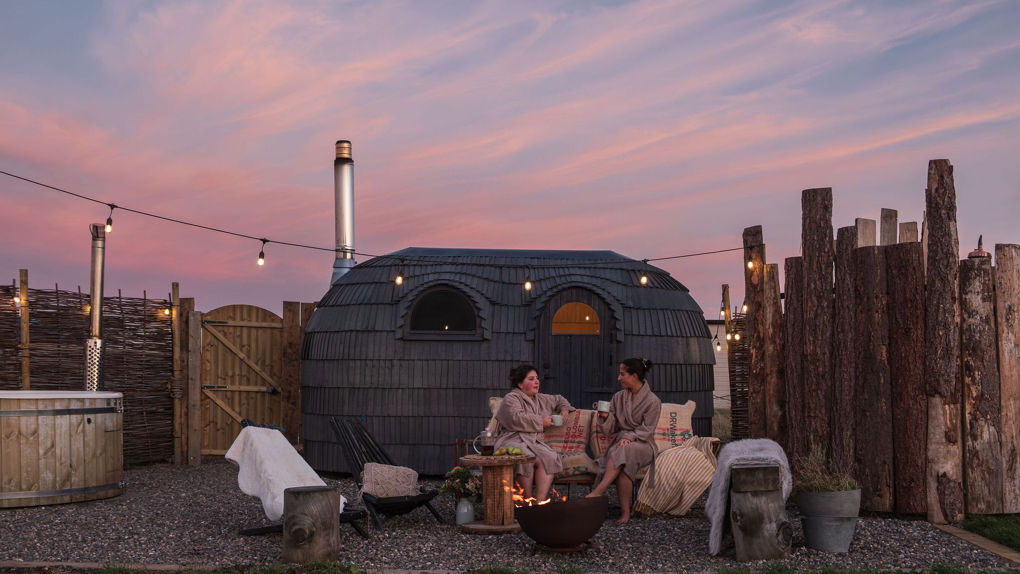 Two people in robes relax by a fire pit at an outdoor spa with a wooden hot tub and a unique Iglusauna  at sunset.