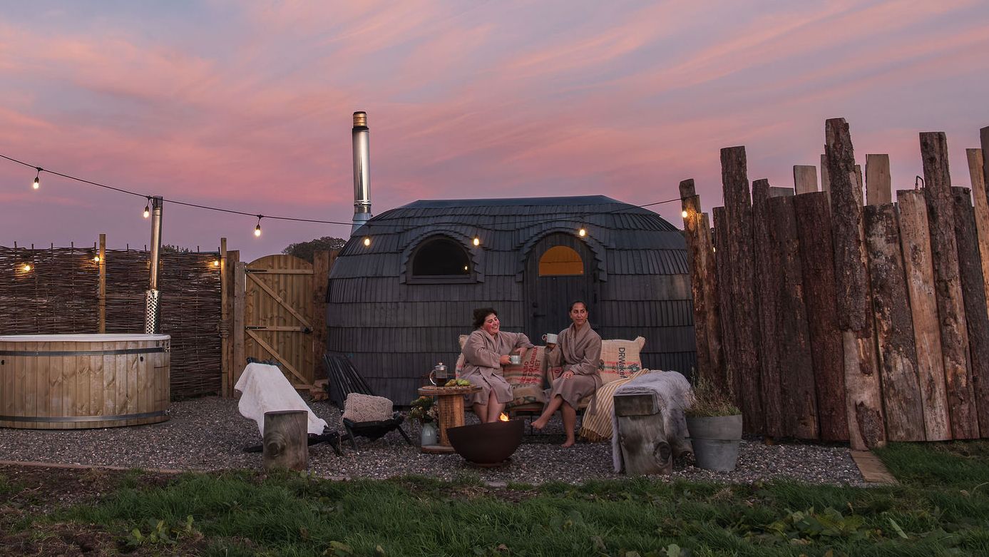 Two people relaxing outside a wooden spa cabin at sunset, surrounded by rustic fencing, a hot tub, and string lights.