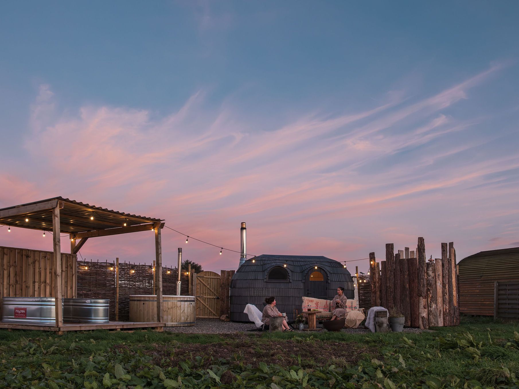 Outdoor wild spa setup at sunset with wooden hot tub, a sauna cabin, and two people relaxing under string lights.