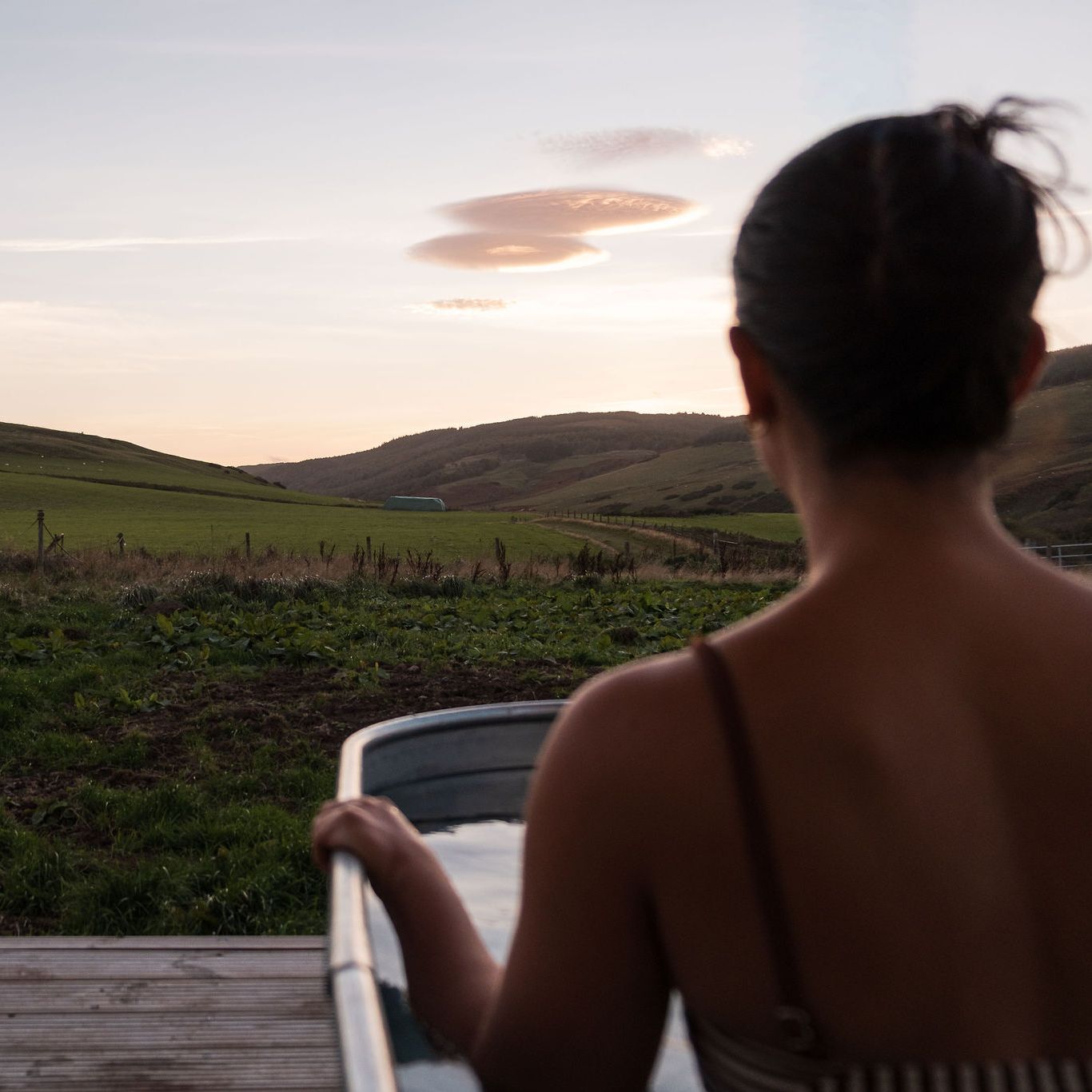 Person relaxing in an outdoor metal spa tub overlooking green hills at sunset.