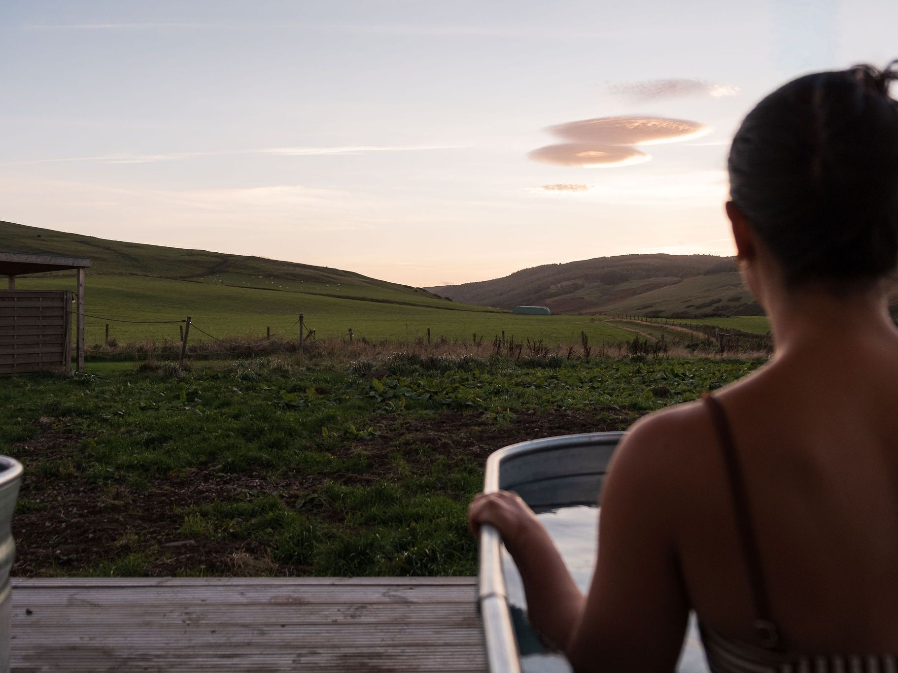 Person relaxing in an outdoor metal spa tub overlooking green hills at sunset.