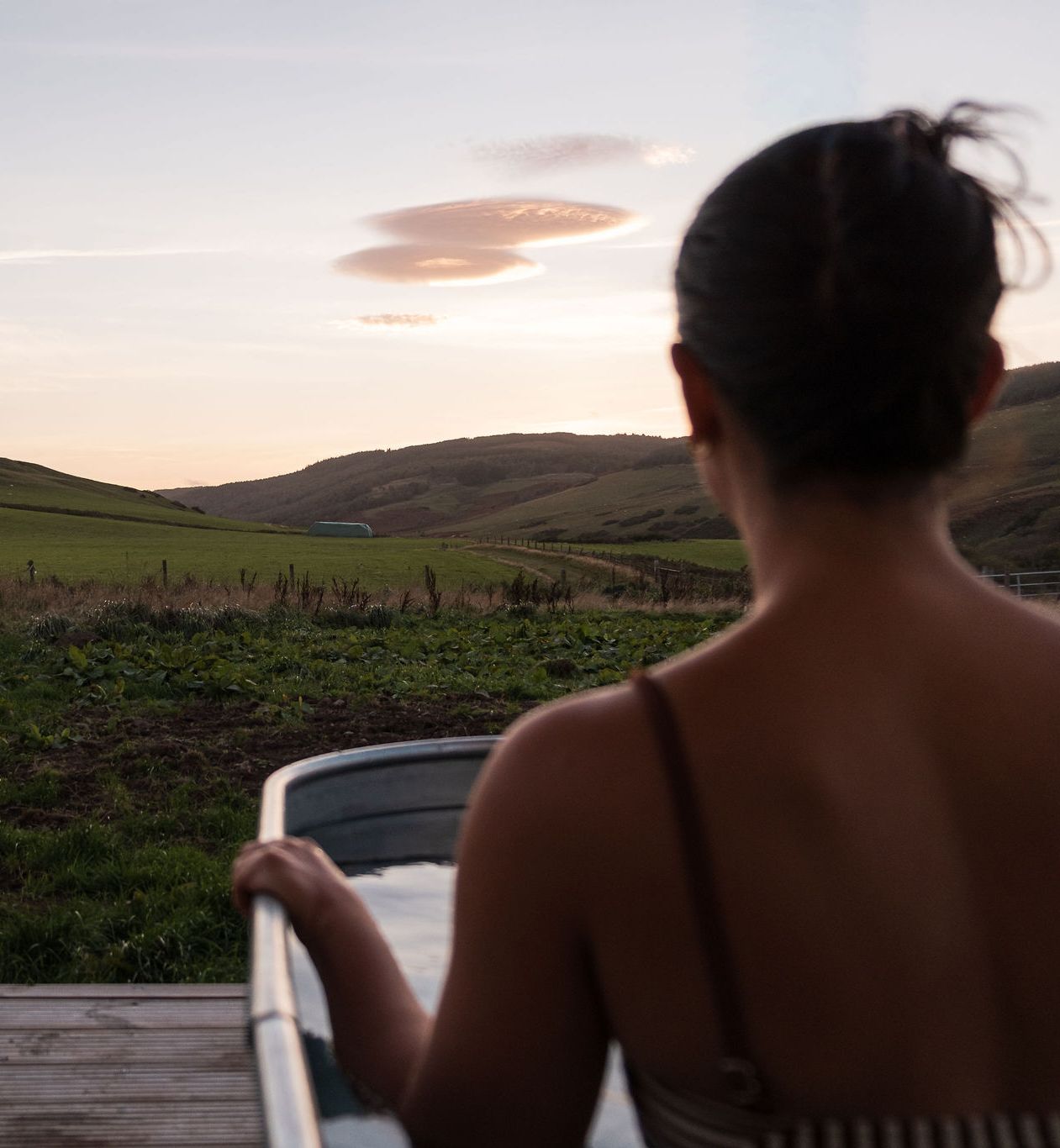 Person relaxing in an outdoor metal spa tub overlooking green hills at sunset.