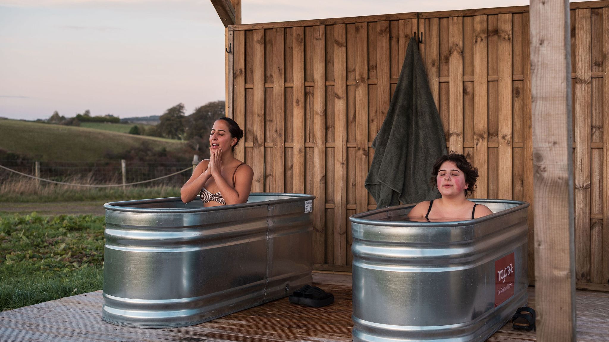 Two women sit in separate metal tubs outdoors, enjoying a cold water spa experience beside a wooden wall and overlooking a scenic landscape.