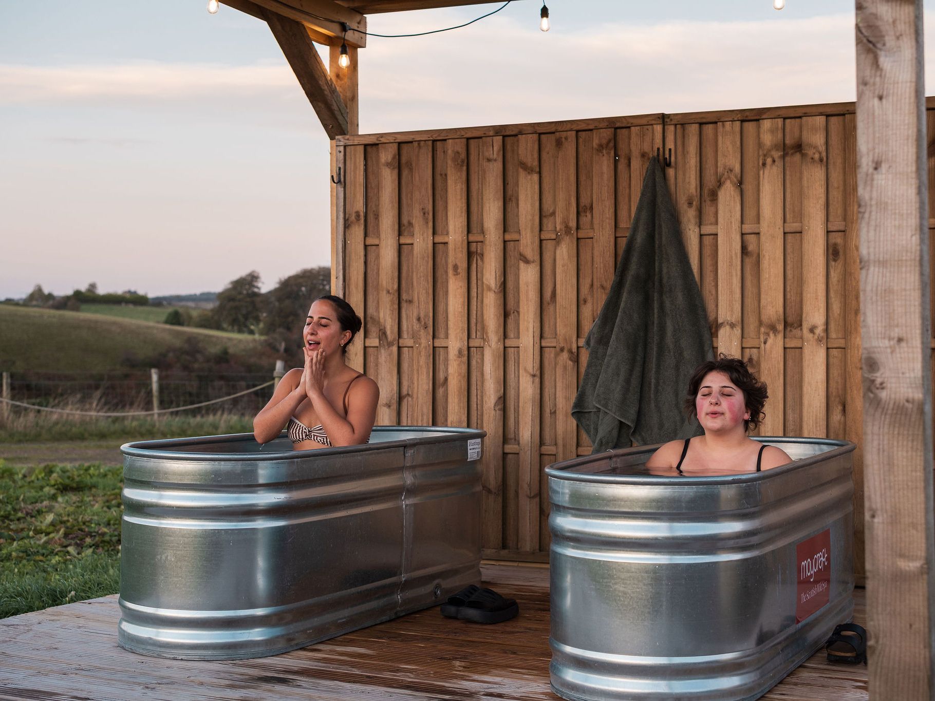 Two women sit in separate metal tubs outdoors, enjoying a cold water spa experience beside a wooden wall and overlooking a scenic landscape.
