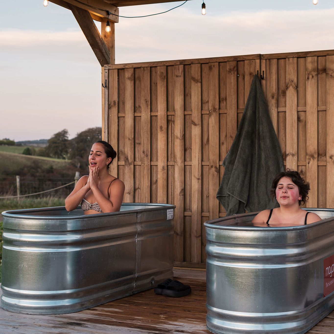 Two women sit in separate metal tubs outdoors, enjoying a cold water spa experience beside a wooden wall and overlooking a scenic landscape.