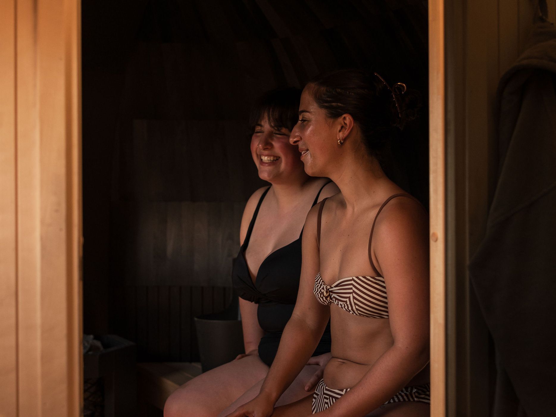 Two women in swimsuits sitting and smiling inside a wooden sauna.