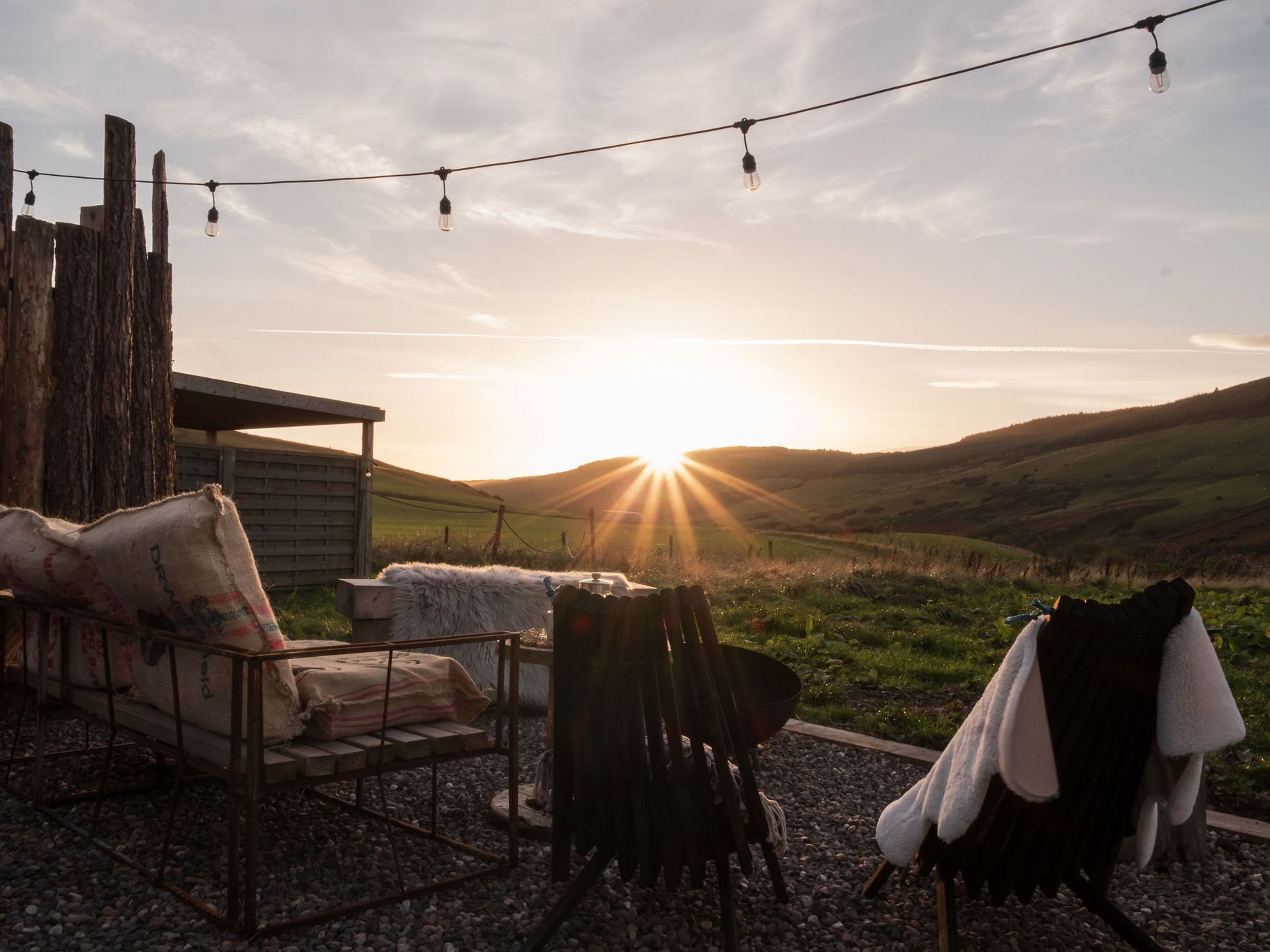 Sunset view from an outdoor seating area with rustic chairs and blankets, overlooking rolling green hills.