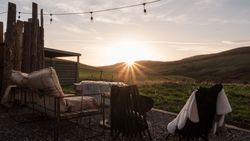 Sunset view from an outdoor seating area with rustic chairs and blankets, overlooking rolling green hills.