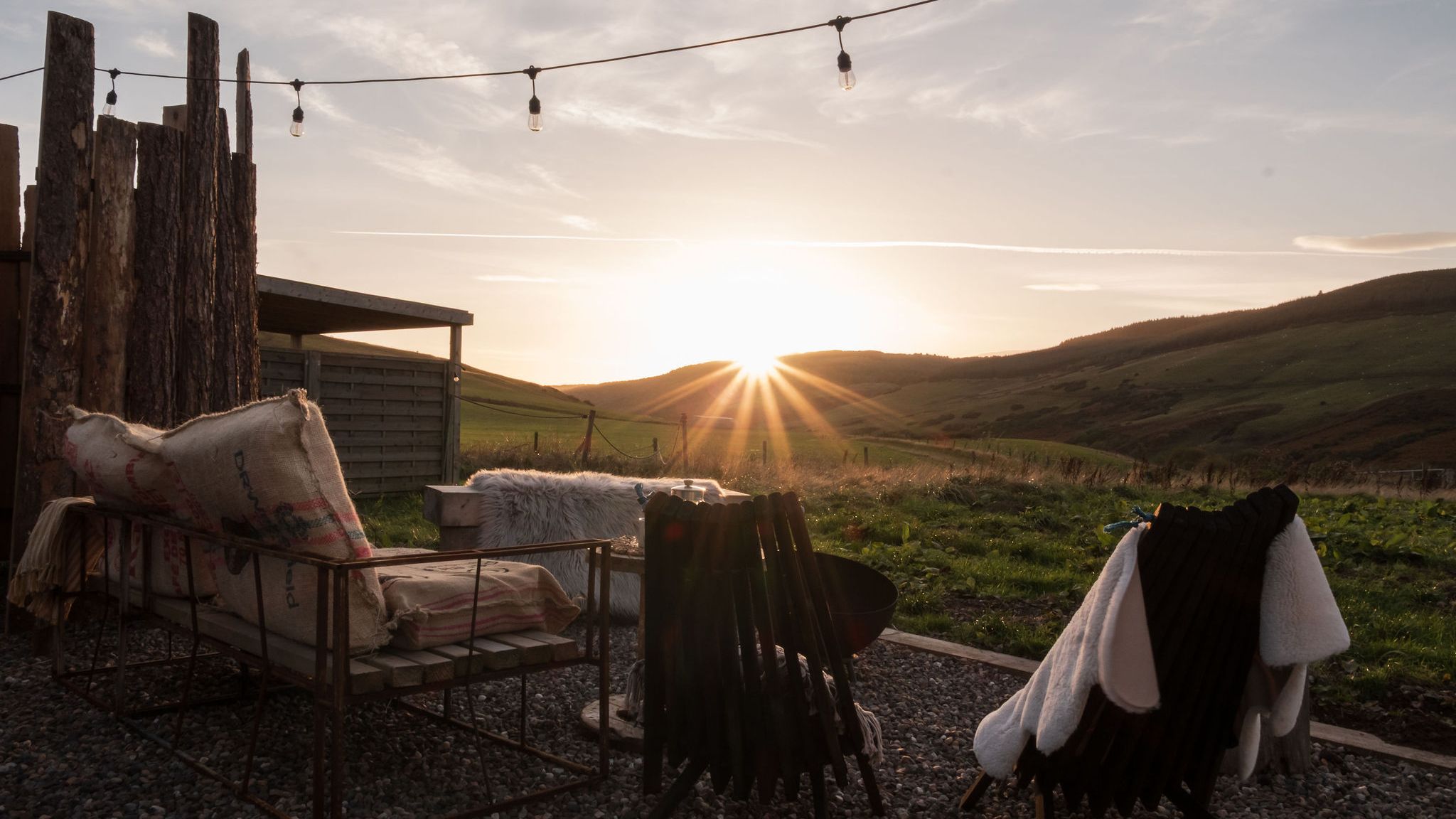 Sunset view from an outdoor seating area with rustic chairs and blankets, overlooking rolling green hills.
