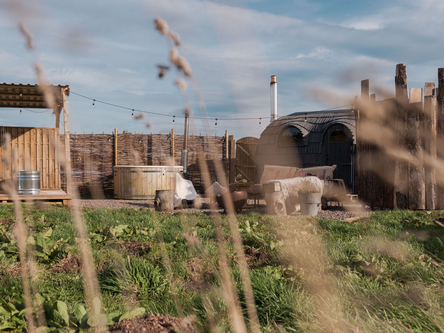 Outdoor wild spa with wooden hot tub, rustic sauna cabin, benches, and natural fencing in a grassy setting.