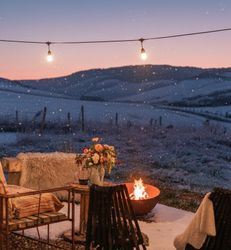 Cozy outdoor seating area with string lights, fire pit, and snow covered hills at sunset