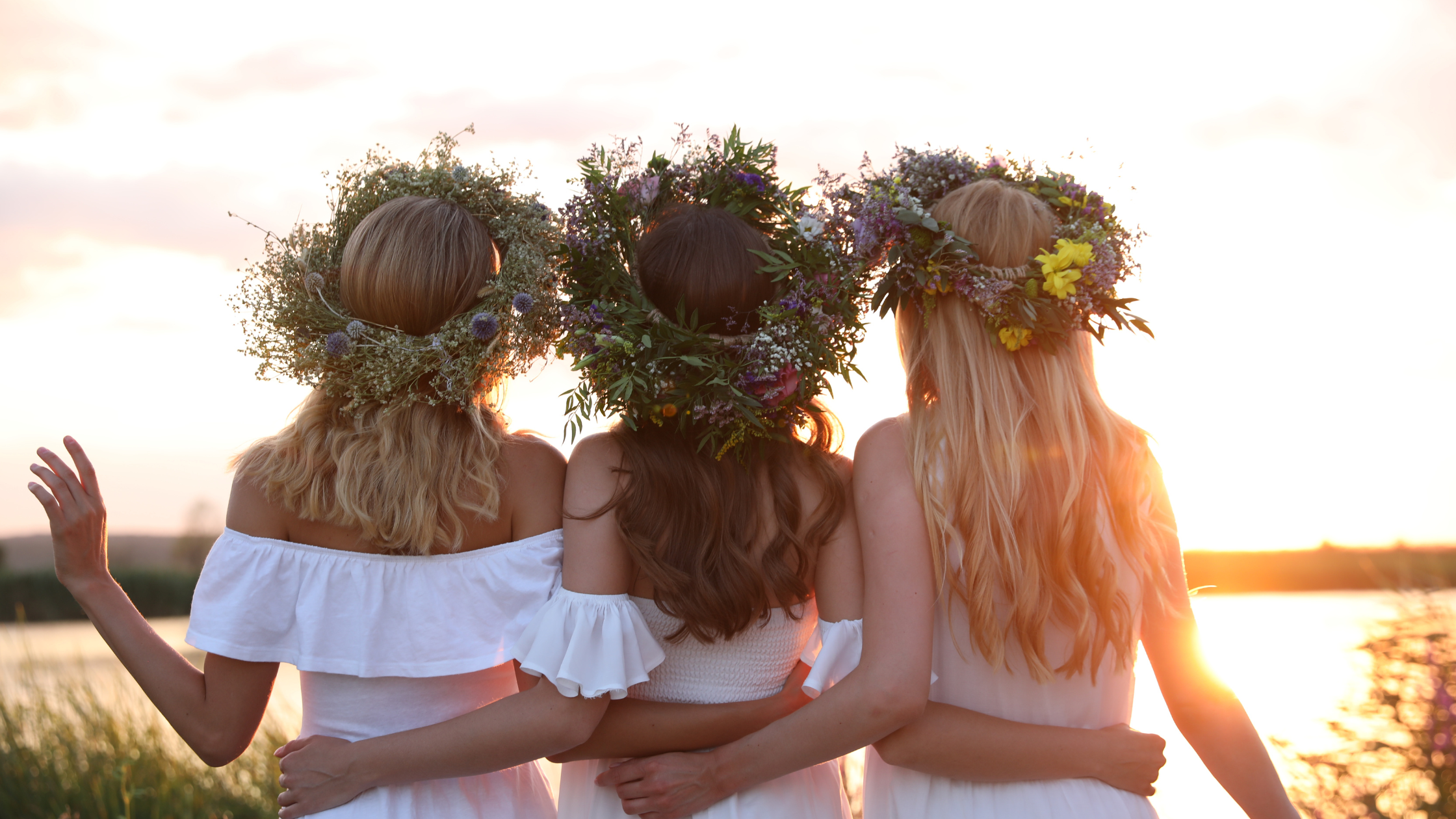 Three women wearing floral wreaths stand with their arms around each other at sunset by a lake.