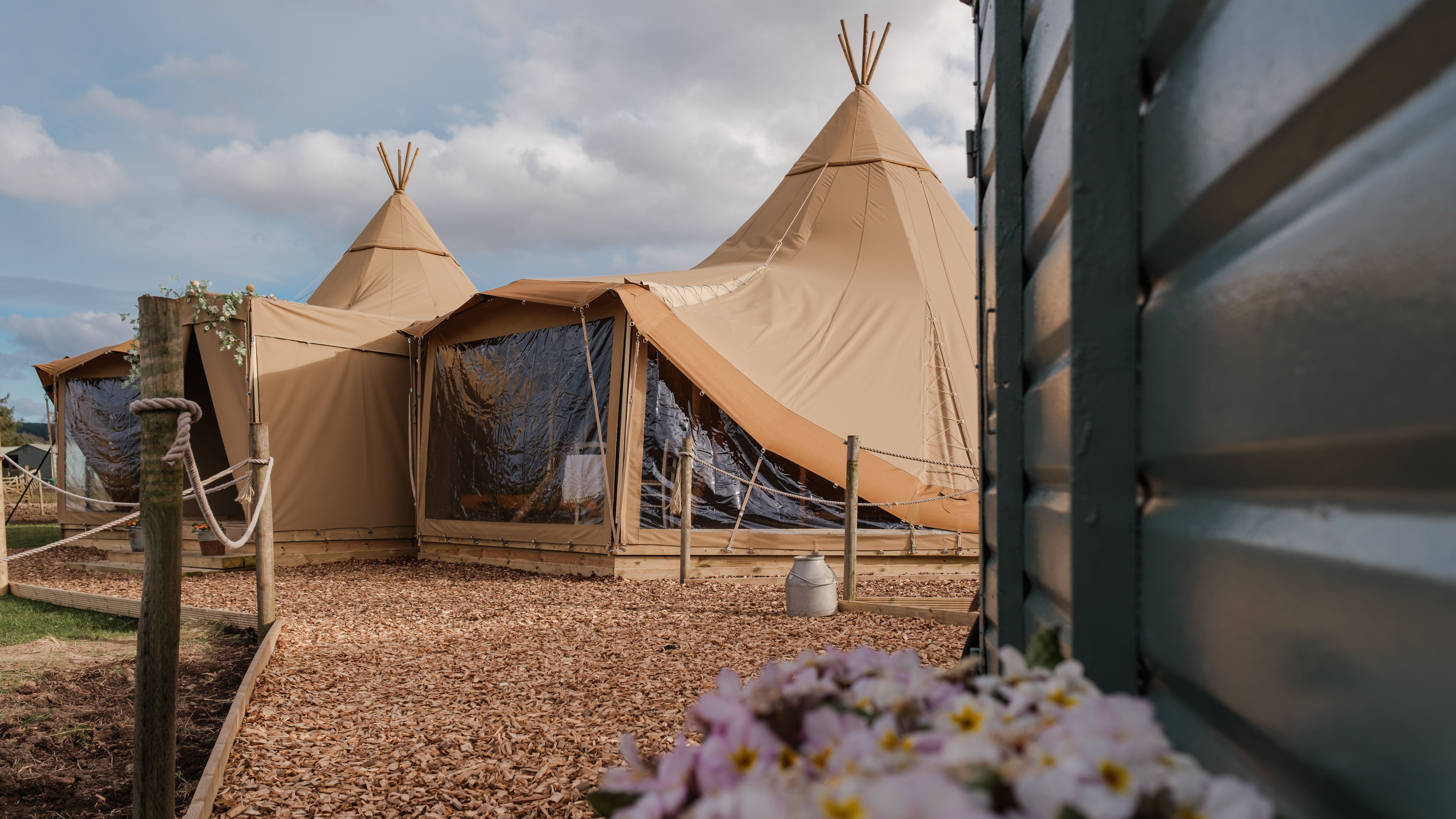 Beige canvas tipi tents with clear window panels beside a wooden cabin under a cloudy sky