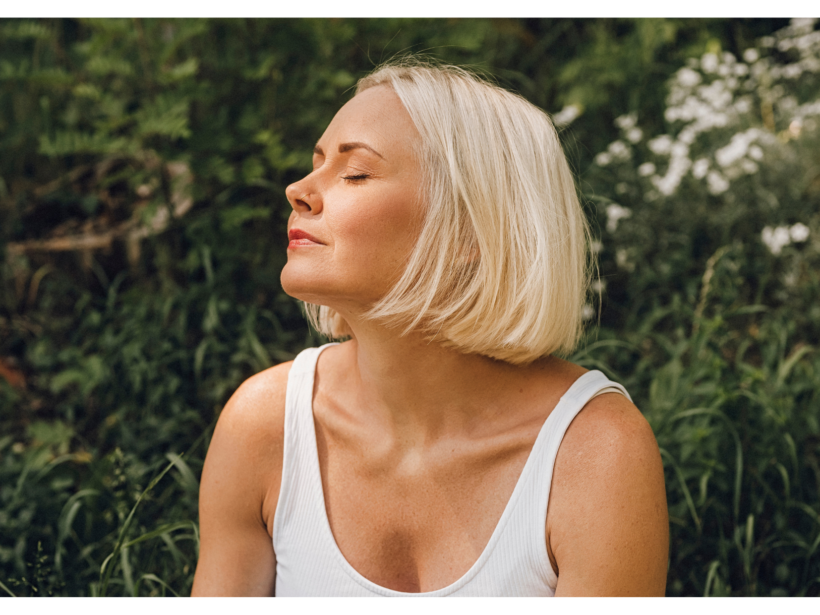 Midlife Blonde woman with eyes closed enjoying sunlight outdoors against a leafy green background