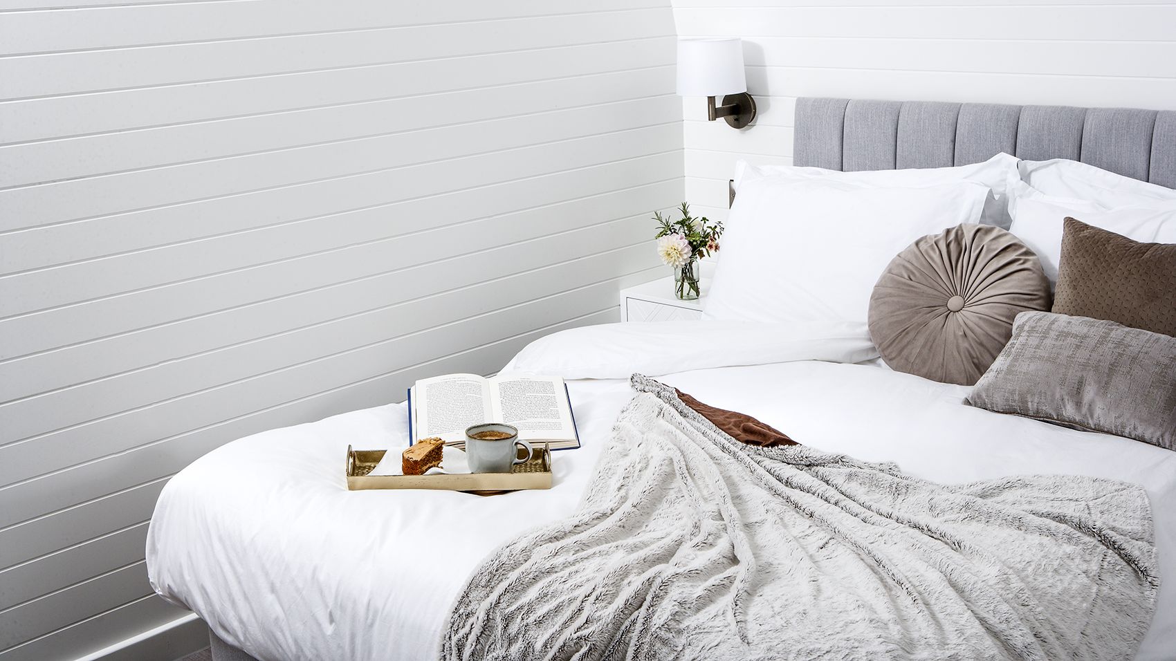 Tray of tea and cake with a book on bed with grey cushions