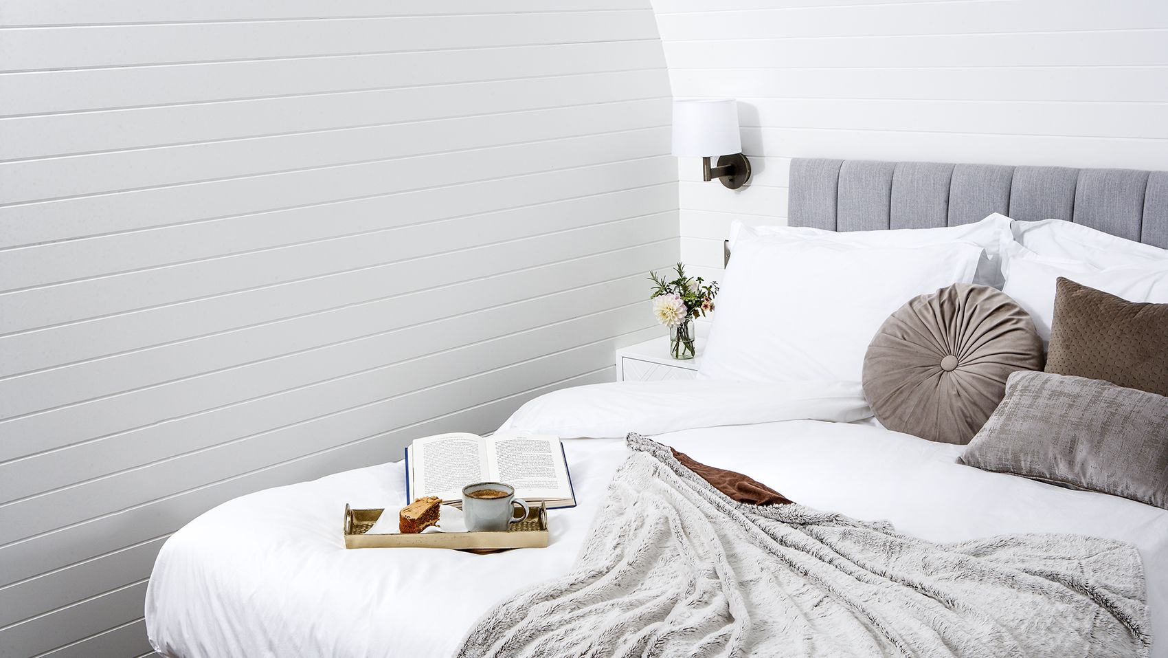 Tray of tea and cake with a book on bed with grey cushions