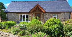 Gwaun Cottage, a stone house with a slate roof and wooden porch, has a cozy patio with seating, surrounded by lush greenery and colourful flowers.