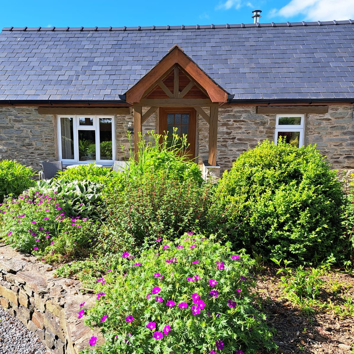 Gwaun Cottage, a stone house with a slate roof and wooden porch, has a cozy patio with seating, surrounded by lush greenery and colourful flowers.