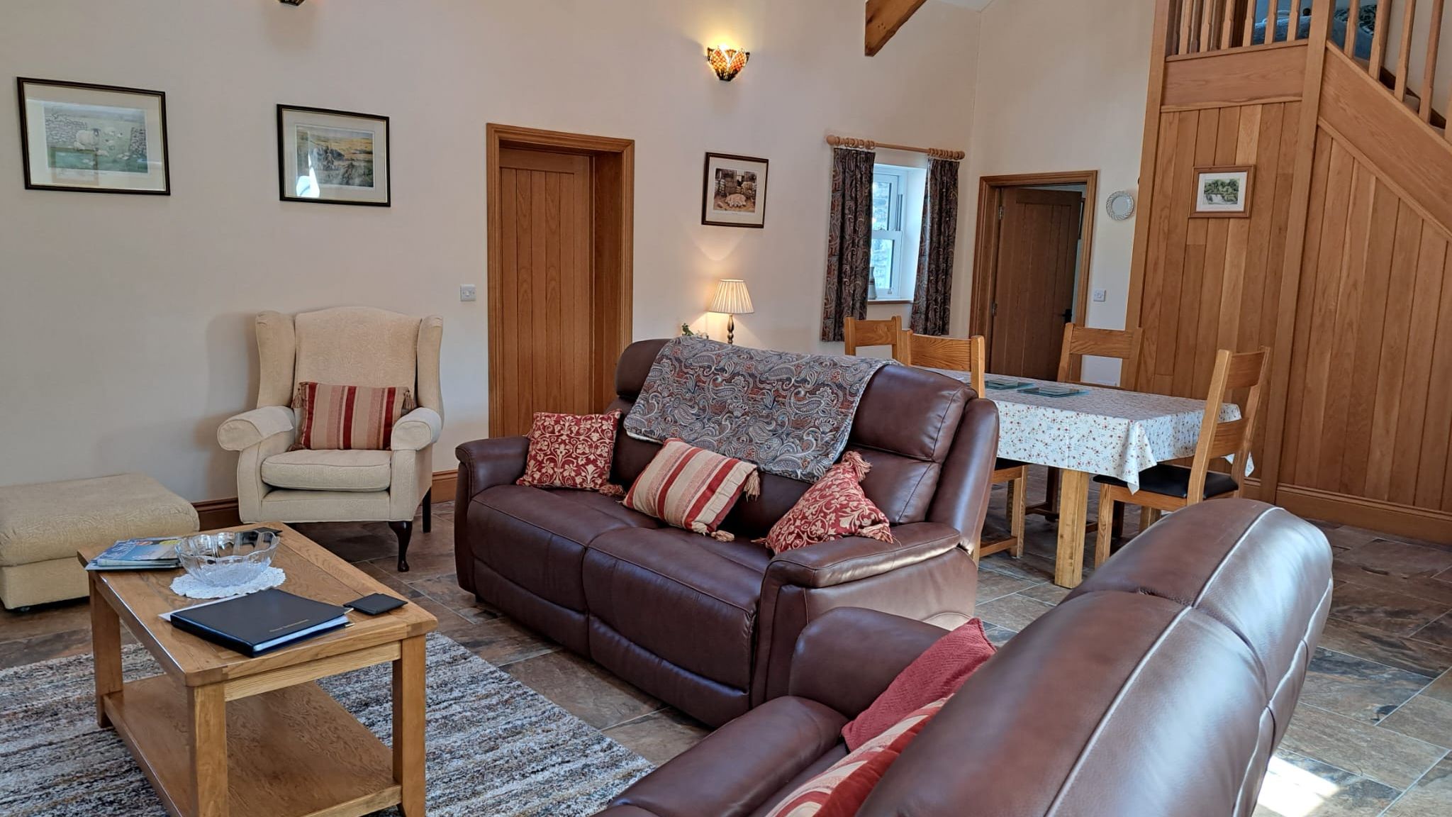 Cozy living room with leather sofas, a beige armchair, wooden coffee table, dining area, and wooden staircase leading upstairs.