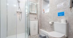 A modern bathroom with grey tiled walls, a curved glass shower enclosure, a white vanity unit with a lit mirror, and a toilet. A small step stool sits near the sink.