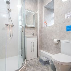 A modern bathroom with grey tiled walls, a curved glass shower enclosure, a white vanity unit with a lit mirror, and a toilet. A small step stool sits near the sink.