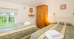 A cosy twin bedroom with two single beds dressed in green and cream bedding. A wooden wardrobe, bedside table with a lamp, and a window with a countryside view complete the space.