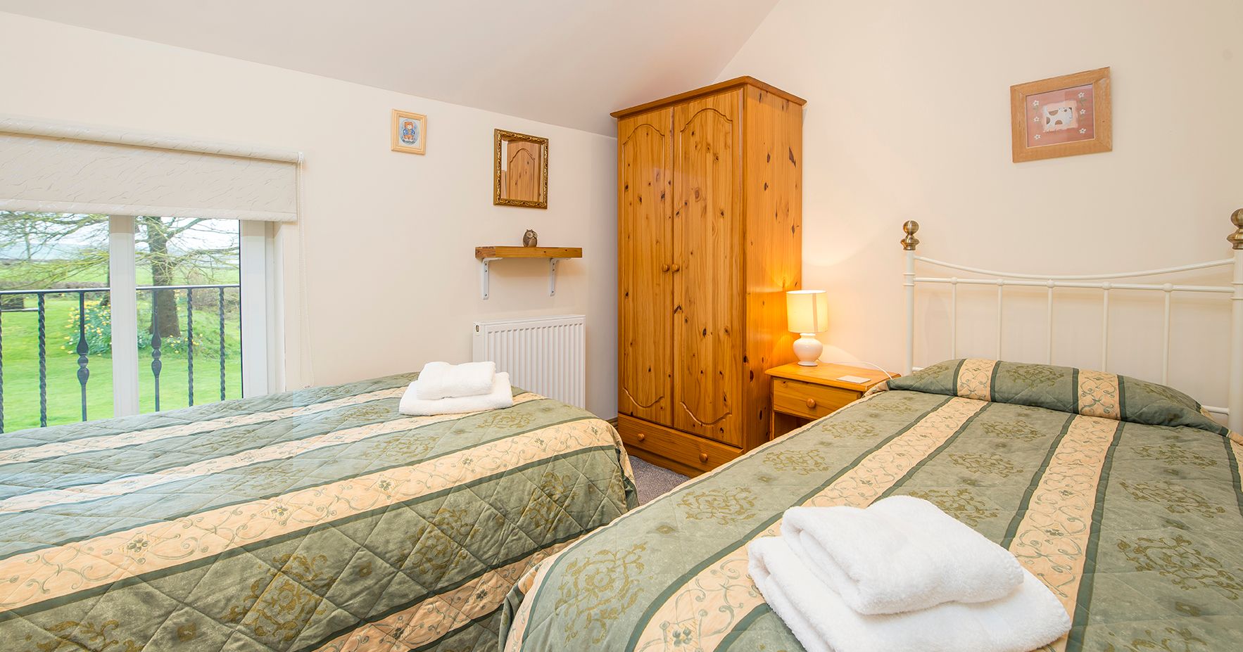 A cosy twin bedroom with two single beds dressed in green and cream bedding. A wooden wardrobe, bedside table with a lamp, and a window with a countryside view complete the space.