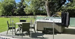 An outdoor patio with a hot tub and a glass-enclosed seating area featuring a round wicker table and chairs. Lush green surroundings and a wooden fence complete the tranquil setting.