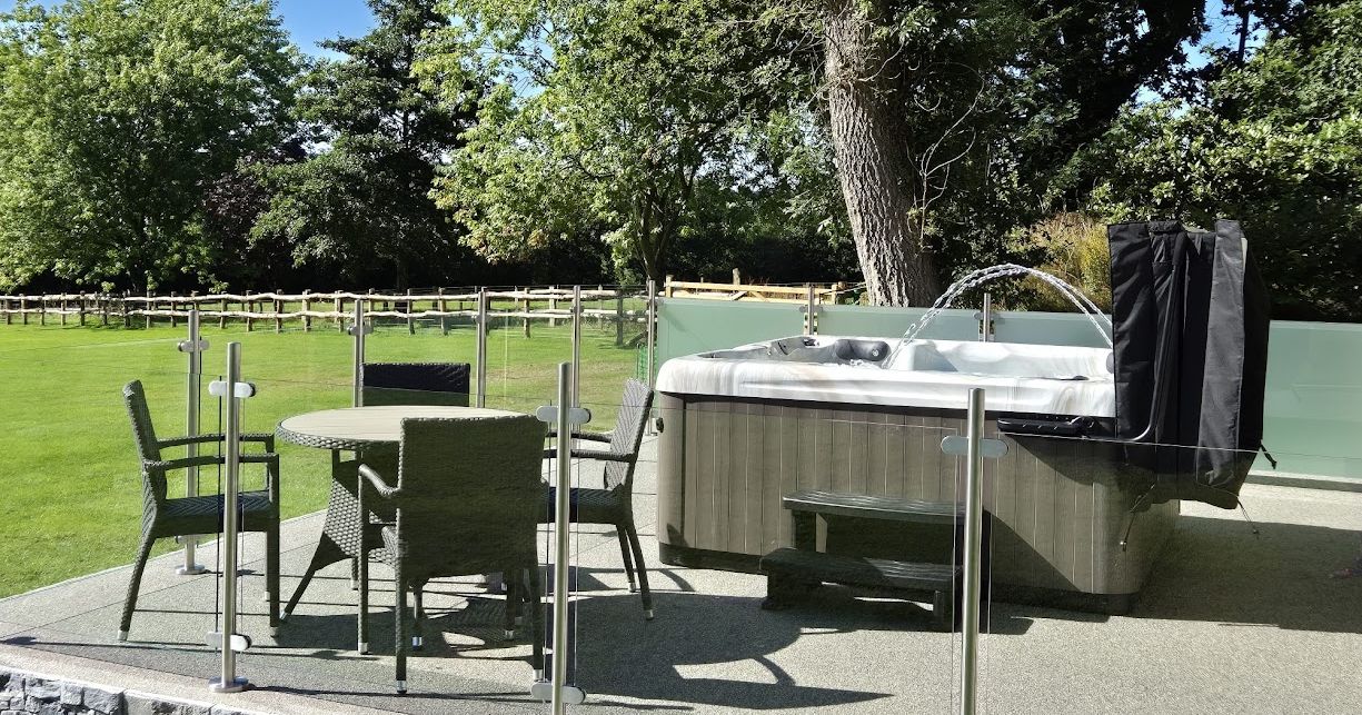 An outdoor patio with a hot tub and a glass-enclosed seating area featuring a round wicker table and chairs. Lush green surroundings and a wooden fence complete the tranquil setting.