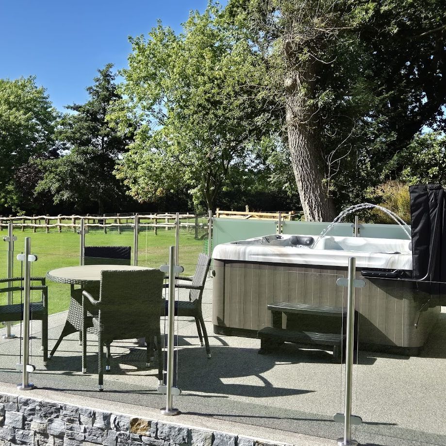 An outdoor patio with a hot tub and a glass-enclosed seating area featuring a round wicker table and chairs. Lush green surroundings and a wooden fence complete the tranquil setting.