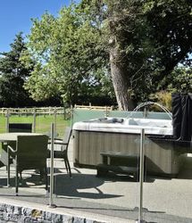 An outdoor patio with a hot tub and a glass-enclosed seating area featuring a round wicker table and chairs. Lush green surroundings and a wooden fence complete the tranquil setting.