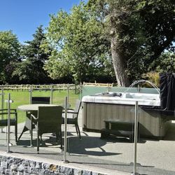 An outdoor patio with a hot tub and a glass-enclosed seating area featuring a round wicker table and chairs. Lush green surroundings and a wooden fence complete the tranquil setting.