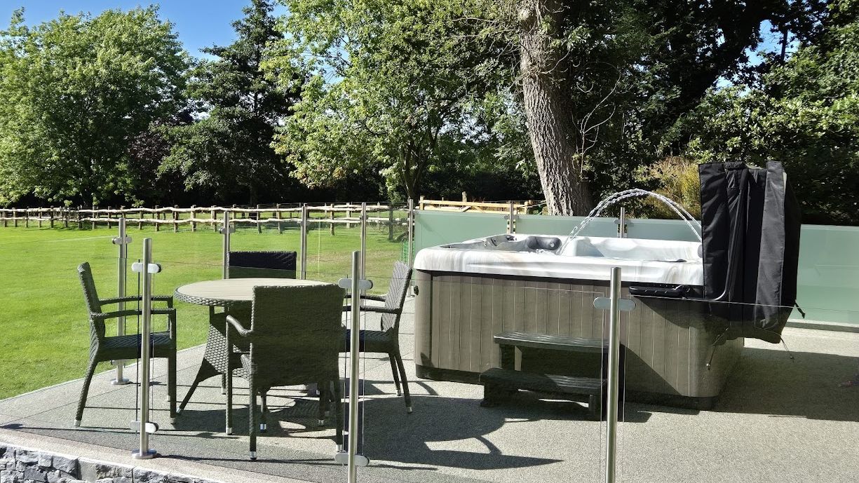 An outdoor patio with a hot tub and a glass-enclosed seating area featuring a round wicker table and chairs. Lush green surroundings and a wooden fence complete the tranquil setting.