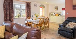 A cosy open-plan living and dining area with a brown leather sofa, a dining table set for four, and a compact kitchen. A stained glass window adds charm to the space.