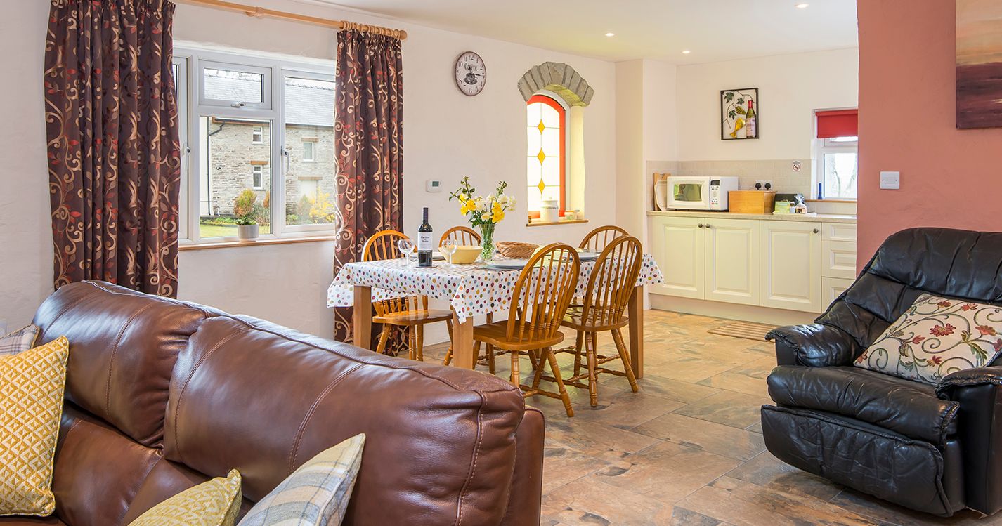 A cosy open-plan living and dining area with a brown leather sofa, a dining table set for four, and a compact kitchen. A stained glass window adds charm to the space.