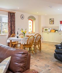 A cosy open-plan living and dining area with a brown leather sofa, a dining table set for four, and a compact kitchen. A stained glass window adds charm to the space.