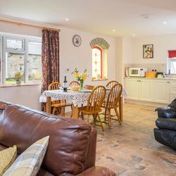 A cosy open-plan living and dining area with a brown leather sofa, a dining table set for four, and a compact kitchen. A stained glass window adds charm to the space.
