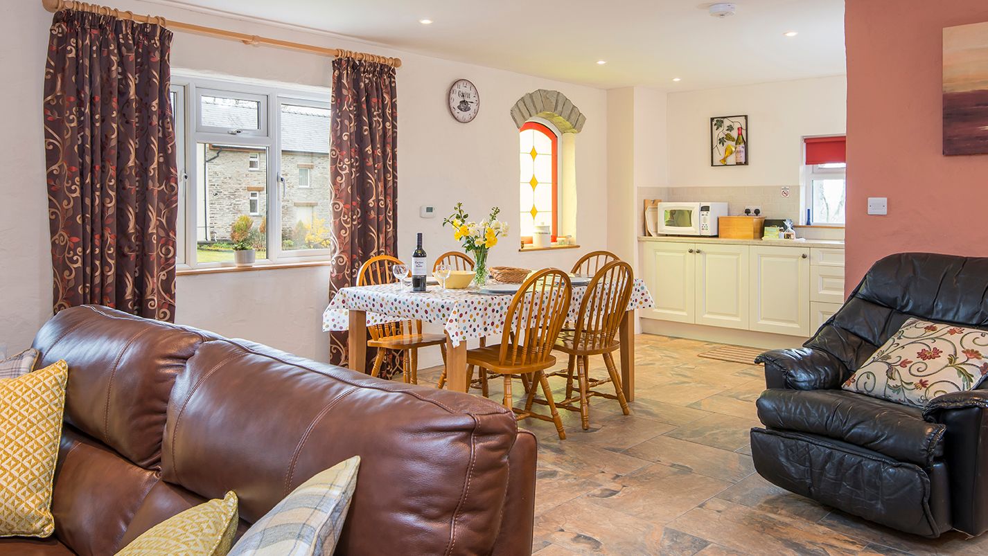 A cosy open-plan living and dining area with a brown leather sofa, a dining table set for four, and a compact kitchen. A stained glass window adds charm to the space.