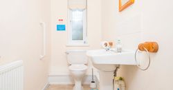 A compact bathroom with a white toilet and pedestal sink. A frosted window lets in natural light, and a grab rail is mounted on the wall for accessibility.