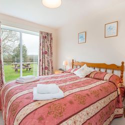 A cosy bedroom with a wooden double bed, red and gold bedding, and matching floral curtains. A sliding glass door opens to a lush green garden with a picnic table.
