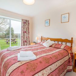 A cosy bedroom with a wooden double bed, red and gold bedding, and matching floral curtains. A sliding glass door opens to a lush green garden with a picnic table.