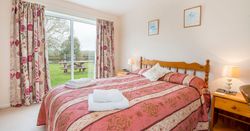A cosy bedroom with a wooden double bed, red and gold bedding, and matching floral curtains. A sliding glass door opens to a lush green garden with a picnic table.