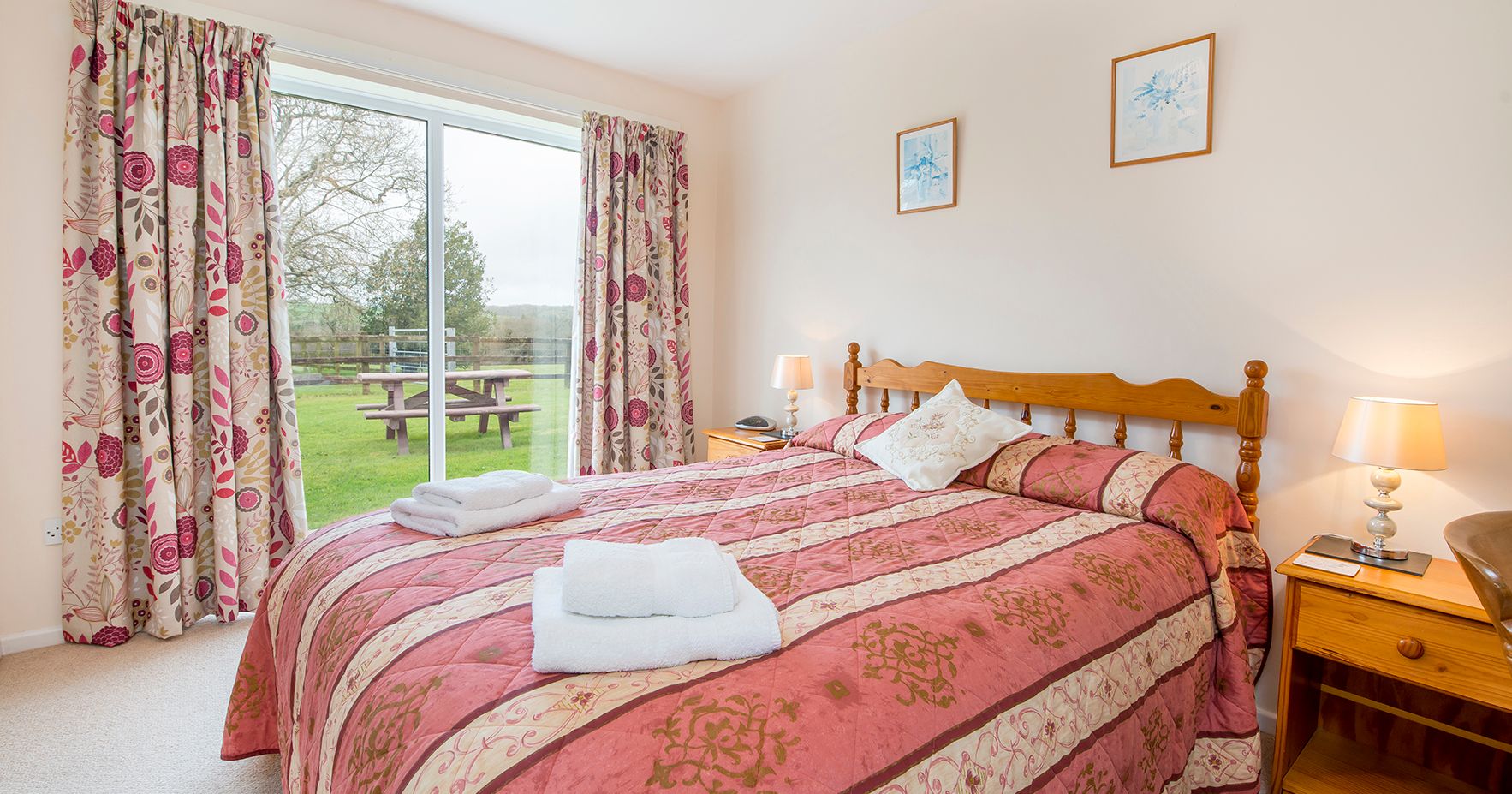 A cosy bedroom with a wooden double bed, red and gold bedding, and matching floral curtains. A sliding glass door opens to a lush green garden with a picnic table.