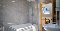 A modern bathroom in Teifi Cottage with a curved bathtub, overhead shower, and sleek grey tiles. A skylight lets in natural light, and an open door leads to the landing.